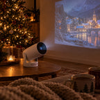 White mini projector on a coffee table showing a snowy village on the wall beside a Christmas tree